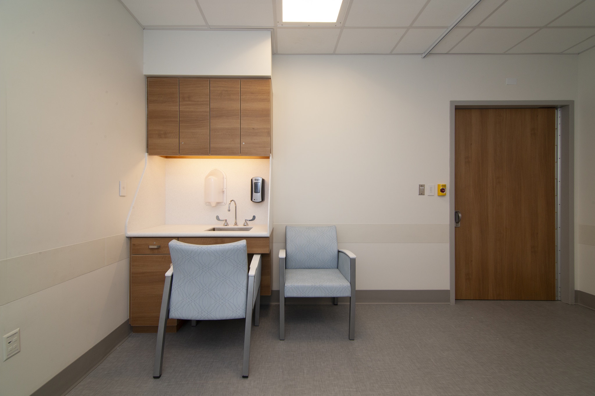 Small exam room with wood cabinetry, sink, light blue chairs, and wood entry door