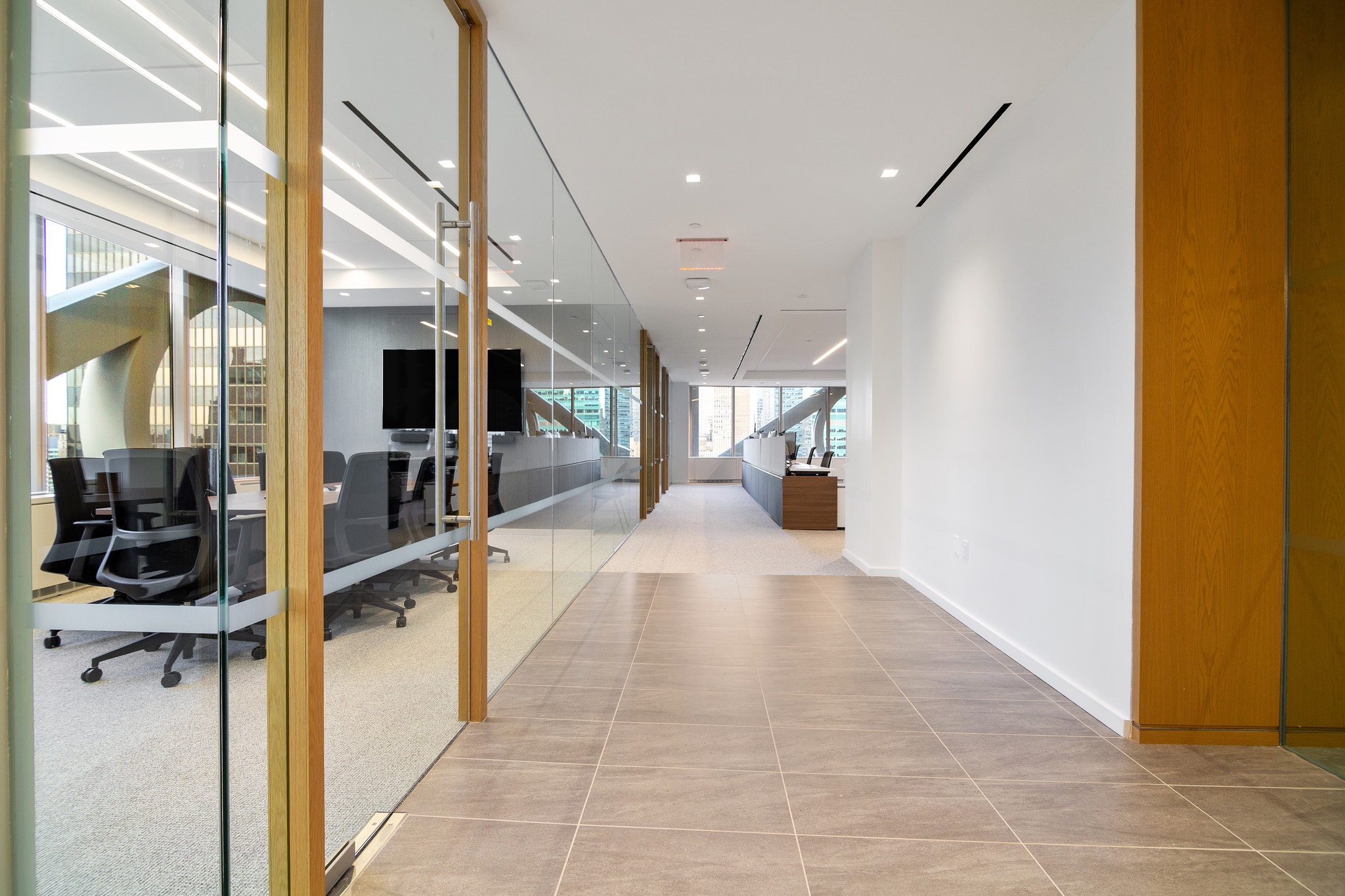 Tiled corridor past glass conference room with black chairs toward wood-paneled reception desk