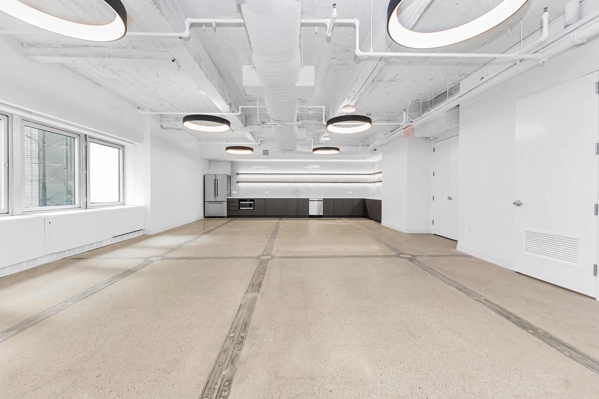 Empty raw office space with exposed white ceiling, round drum pendants, and gray kitchenette against back wall