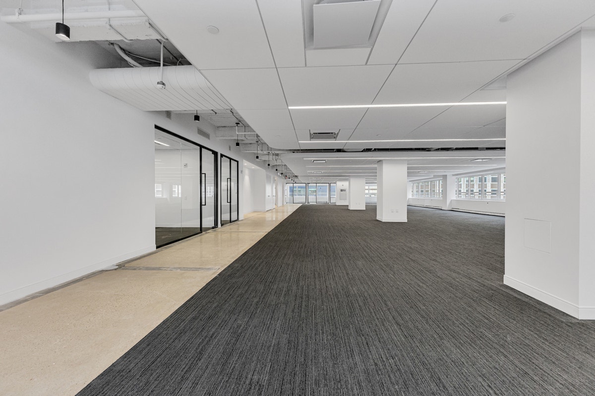 Long open office corridor with dark carpet, glass-front conference rooms, and exposed ceiling ducts
