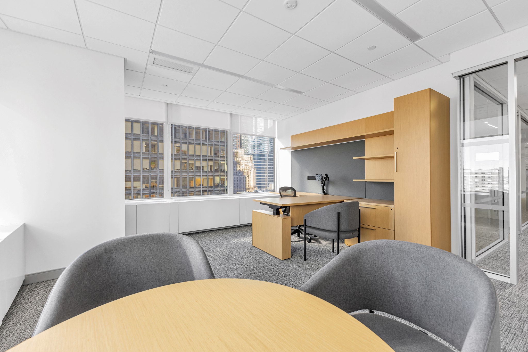 Corner office with wood desk, gray guest chairs, round conference table, and city skyline views through floor-to-ceiling windows
