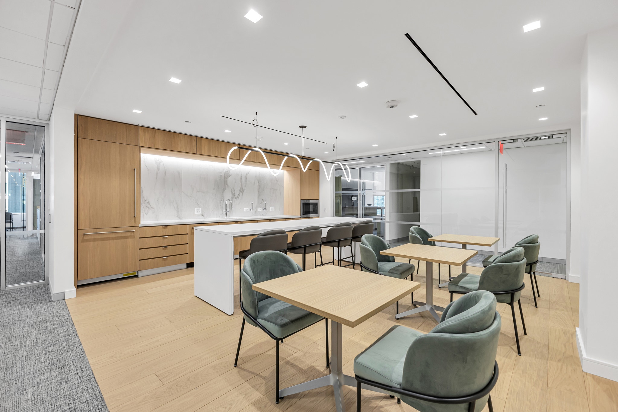 Modern break room with sculptural pendant light, marble backsplash kitchen, and green velvet chairs at wood tables