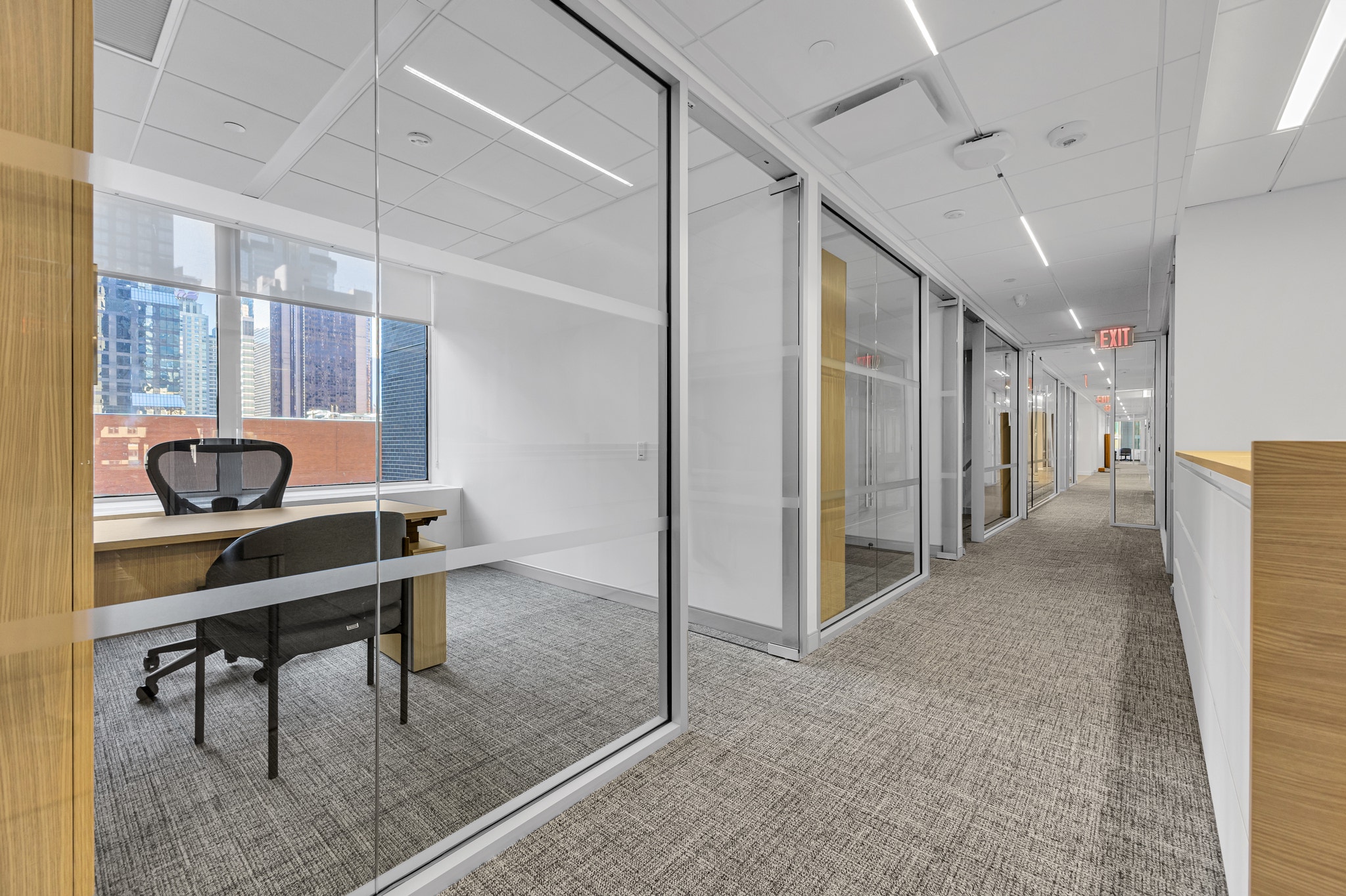 Glass-walled private offices along a carpeted corridor with city views and illuminated exit sign