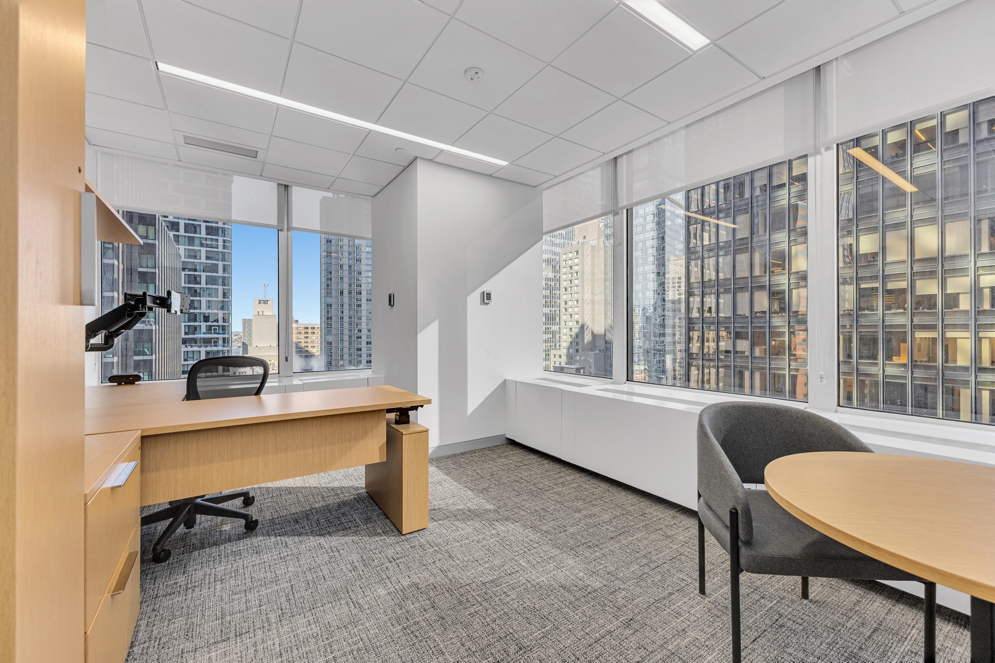 Private office corner with wood desk, monitor arm, round meeting table, and wraparound skyscraper views