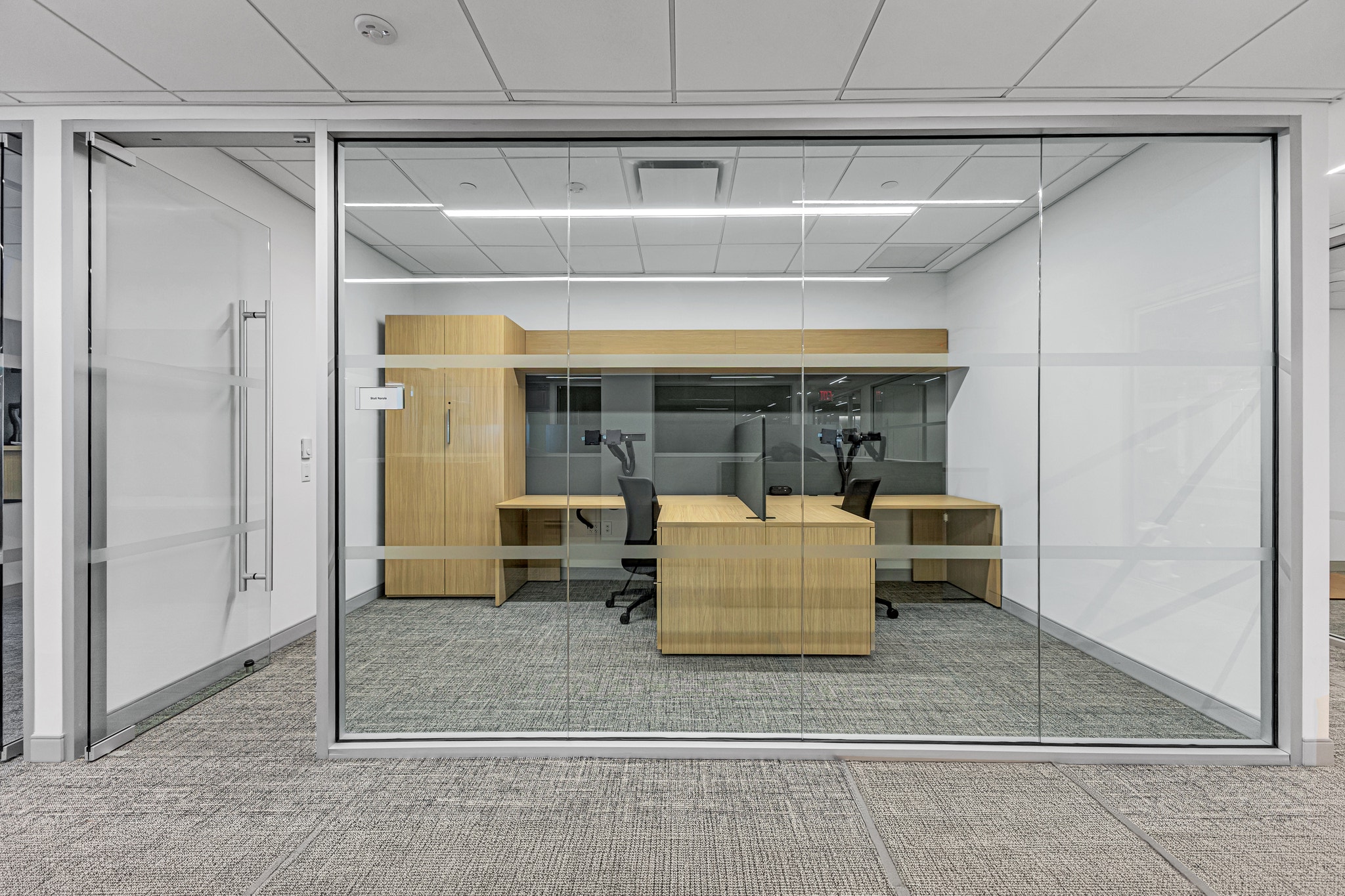 Glass-front shared office with two wood desks, privacy screen, tall wood storage cabinet, and task chairs